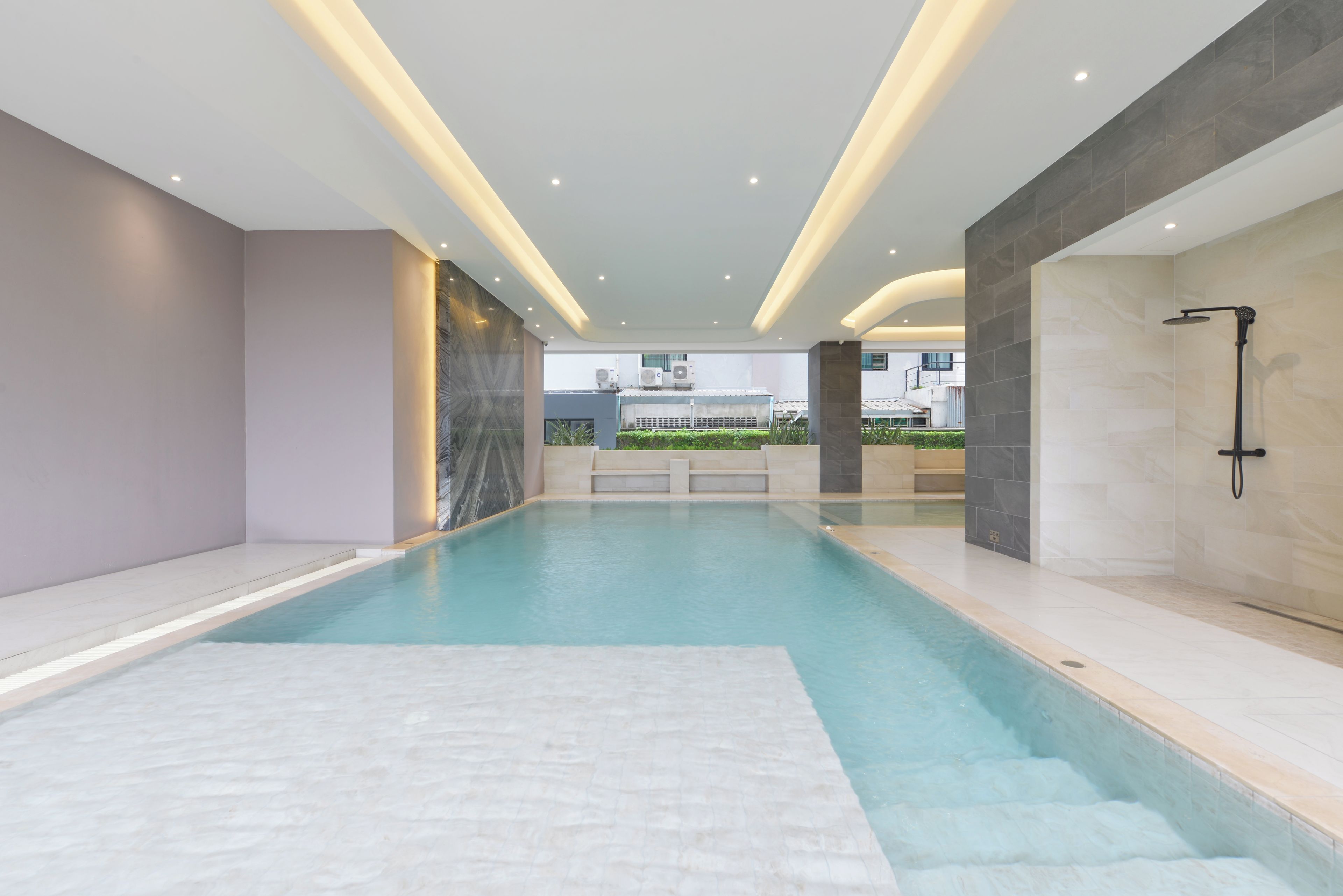 Una piscina interior moderna con vistas despejadas del entorno, que incluye un edificio con unidades de aire acondicionado y un jardín ajardinado.