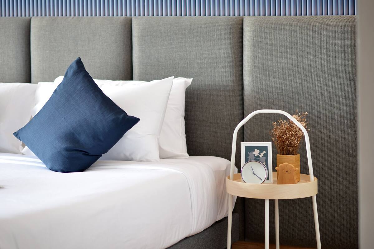 A neatly made bed with white and blue pillows, a bedside table with a clock, a framed picture, and a vase with dried flowers.