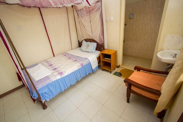 Un dormitorio con una cama individual, una mesita de noche de madera, una silla, un mueble de madera pequeño y un baño con lavabo y espejo.