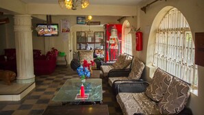 A lobby with checkered flooring, a glass coffee table with a red candle, patterned sofas, a television, and a Coca-Cola refrigerator. - Heritage Bliss Hotel (Kitui)