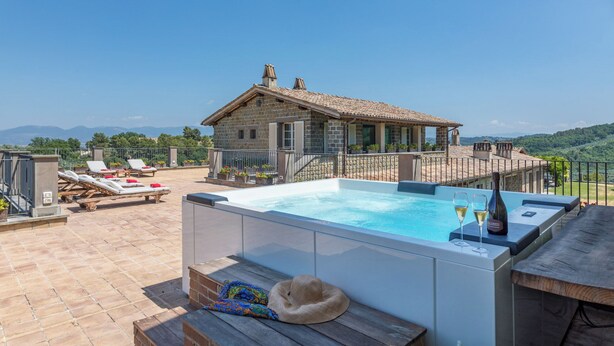 Zona d'una piscina al terrat amb gandules, una banyera d'hidromassatge i vista cap a una casa de pedra amb teulada aïllada.