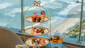 A three-tiered dessert stand with various sweets and a teapot on a table with a view of the ocean. - Rosamia Da Nang Hotel (Da Nang)