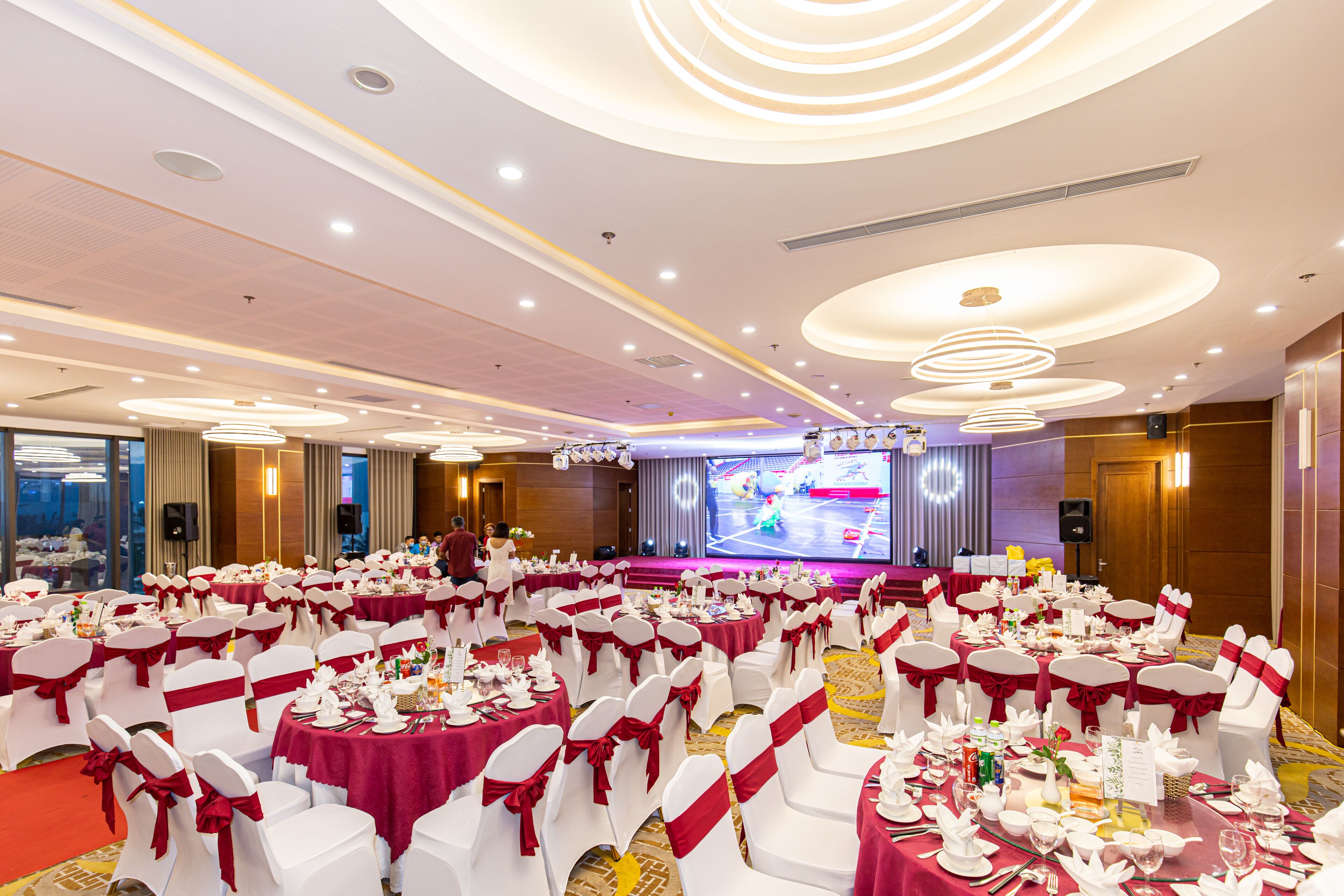 A banquet hall with round tables, white chairs, and red sashes, featuring a stage and a large screen in the background.