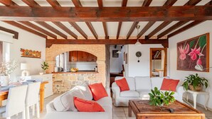 Un'ampia zona giorno con camino in pietra, divani bianchi con cuscini rossi, un tavolino da caffè in legno e una cucina visibile attraverso un arco.