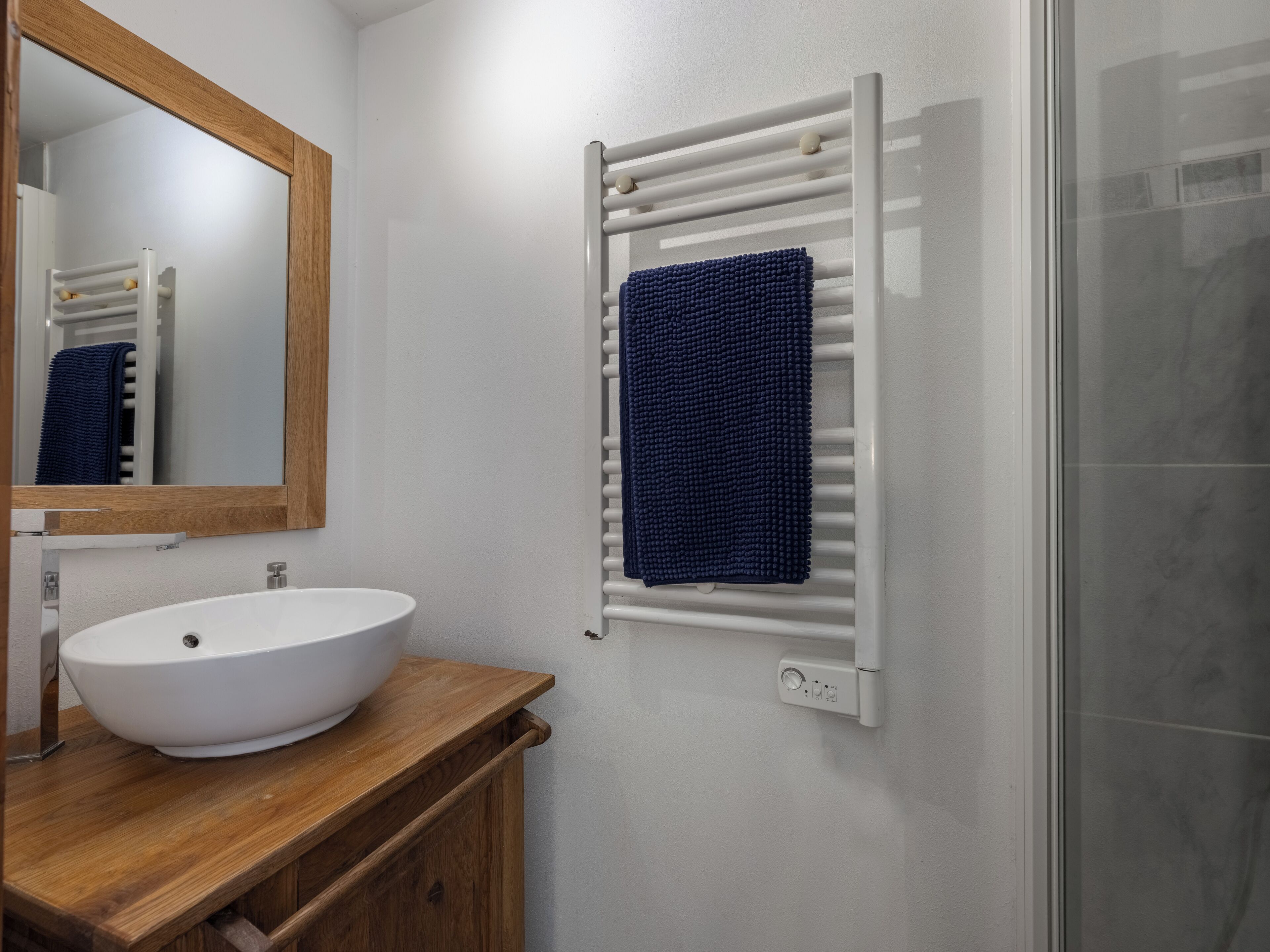 Een badkamer met een witte wastafel, houten opbergmeubel, een spiegel, een handdoekhouder met een blauwe handdoek en een douchecabine met glazen deur.