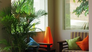 Ein Zimmer mit einer Zimmerpflanze, einem Tisch, einem Sessel und einer Couch mit Kissen. Zwei Fenster mit Rundbögen sind vorhanden.