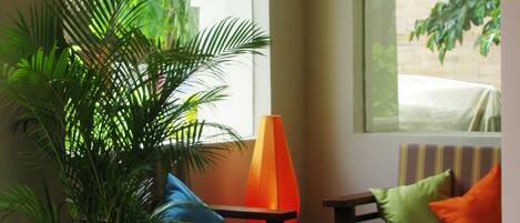 A room with a potted plant, a table, a chair, and a sofa with cushions. There are two windows with arched tops.
