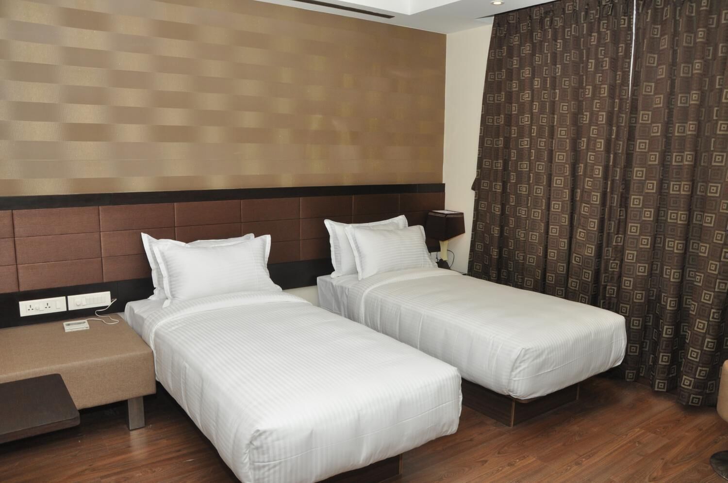 A hotel room with two single beds, a nightstand, a lamp, and a patterned curtain.