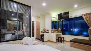 A hotel room with a large bed, a desk, a TV, and a window with a view of the night sky. - Pura Nakhon Hotel (Nakhon Si Thammarat)