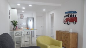 Una sala moderna con un sofá, un sillón amarillo, un mueble de madera y una cocina con refrigerador, lavamanos y mesa de comedor.