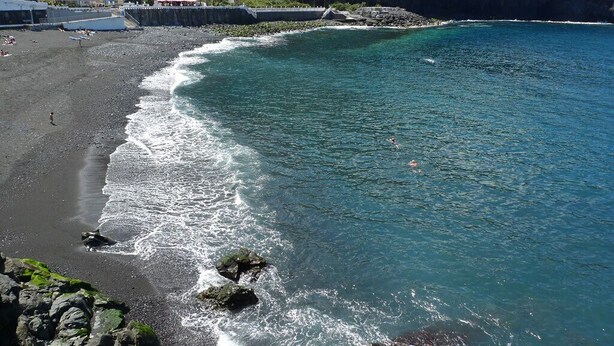 Una playa con agua azul cristalina, costa rocosa y edificios al fondo.