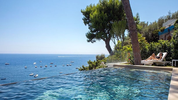 A pool with clear blue water, a view of the sea with boats, and a person relaxing on a lounge chair. - AMORE RENTALS - Villa Ibiscus with Infinity Pool, Direct Sea Access, Sea View, Parking and Breakfast (Nerano)