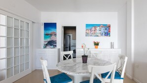 Een eetruimte met een ronde marmeren tafel, witte stoelen met blauwe kussens en twee schilderijen aan de muur.