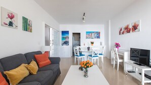 Een moderne woonkamer met een grijze bank, een witte salontafel en een televisie op een witte dressoir. Achterin staan twee eetkamerstoelen en een tafel.