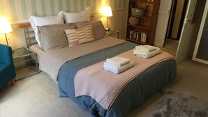 A neatly made bed with pillows, a bedside table with a lamp, a bookshelf, and a fluffy rug on the floor. - HOWTON COTTAGE - village cottage on coast path next to beaches (Cawsand)