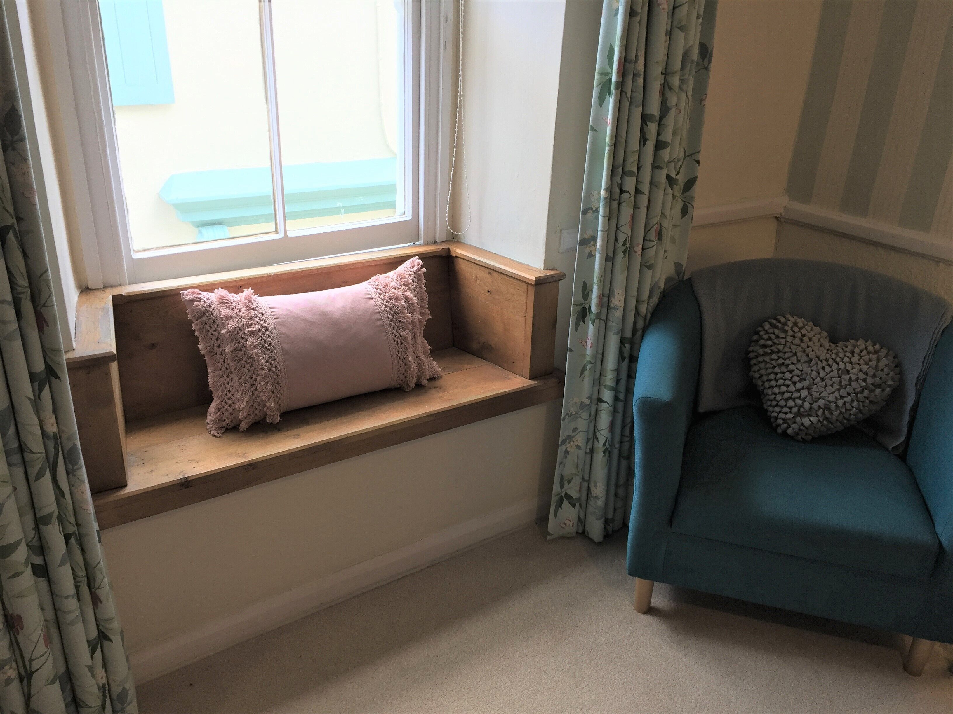 Ein Zimmer mit Fensterbank, blauem Sessel und Herz-Kissen.