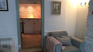 A cozy room with a light blue armchair, a framed picture on the wall, and a kitchen visible through an open doorway. - HOWTON COTTAGE - village cottage on coast path next to beaches (Cawsand)