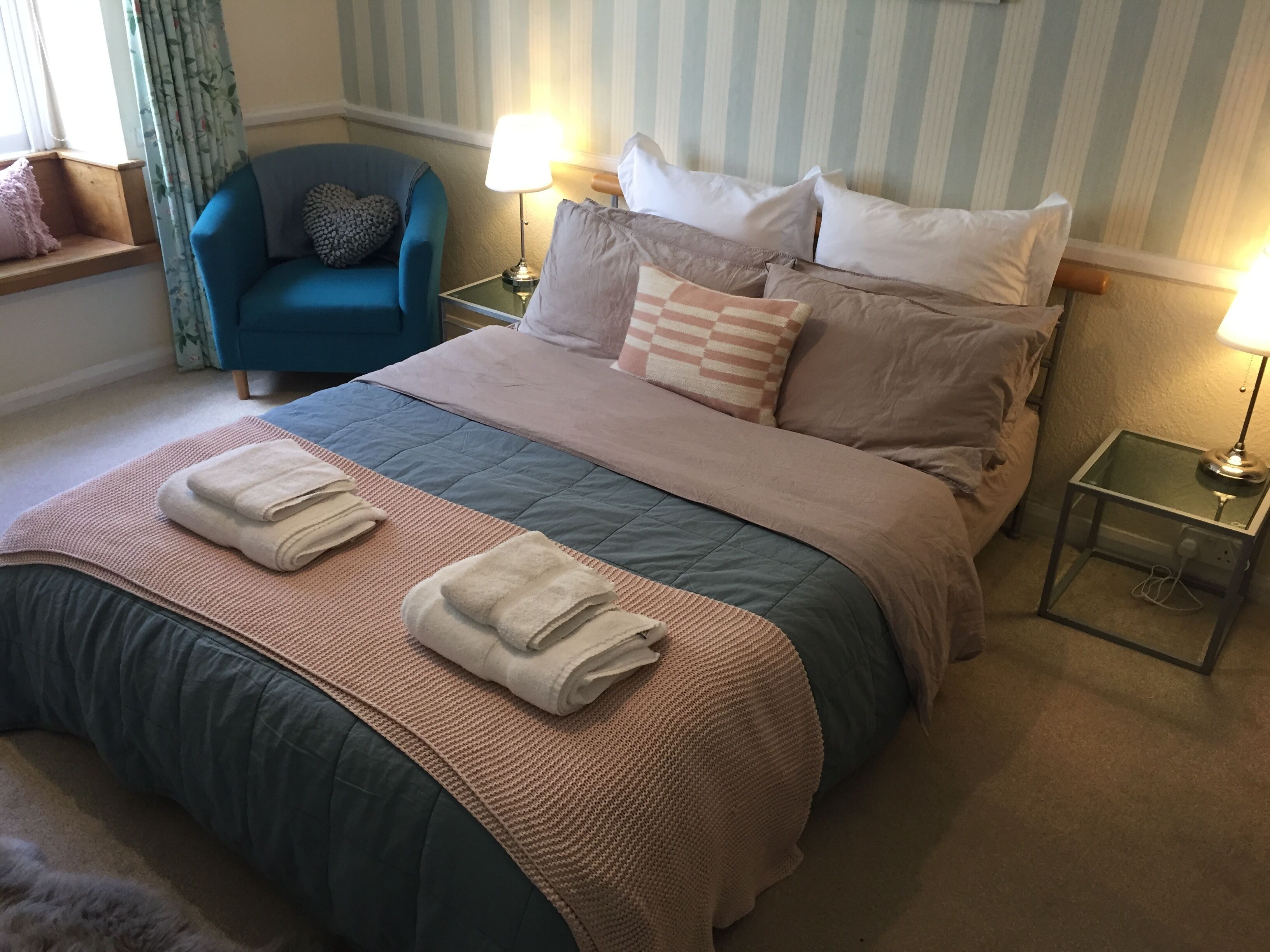 A neatly made bed with a pink and blue quilt, two bedside lamps, a blue armchair, and a window with curtains.