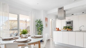 Une cuisine moderne avec des meubles blancs, un four à micro-ondes et un évier. On y trouve également un coin repas avec une table en verre et des chaises blanches.