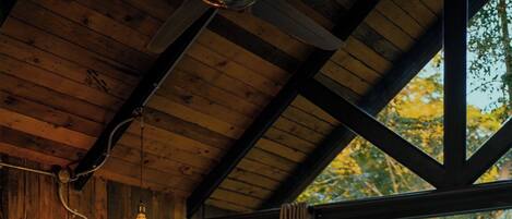 A wooden cabin bedroom with a bed, a nightstand, a lamp, and a view of trees through a large window.