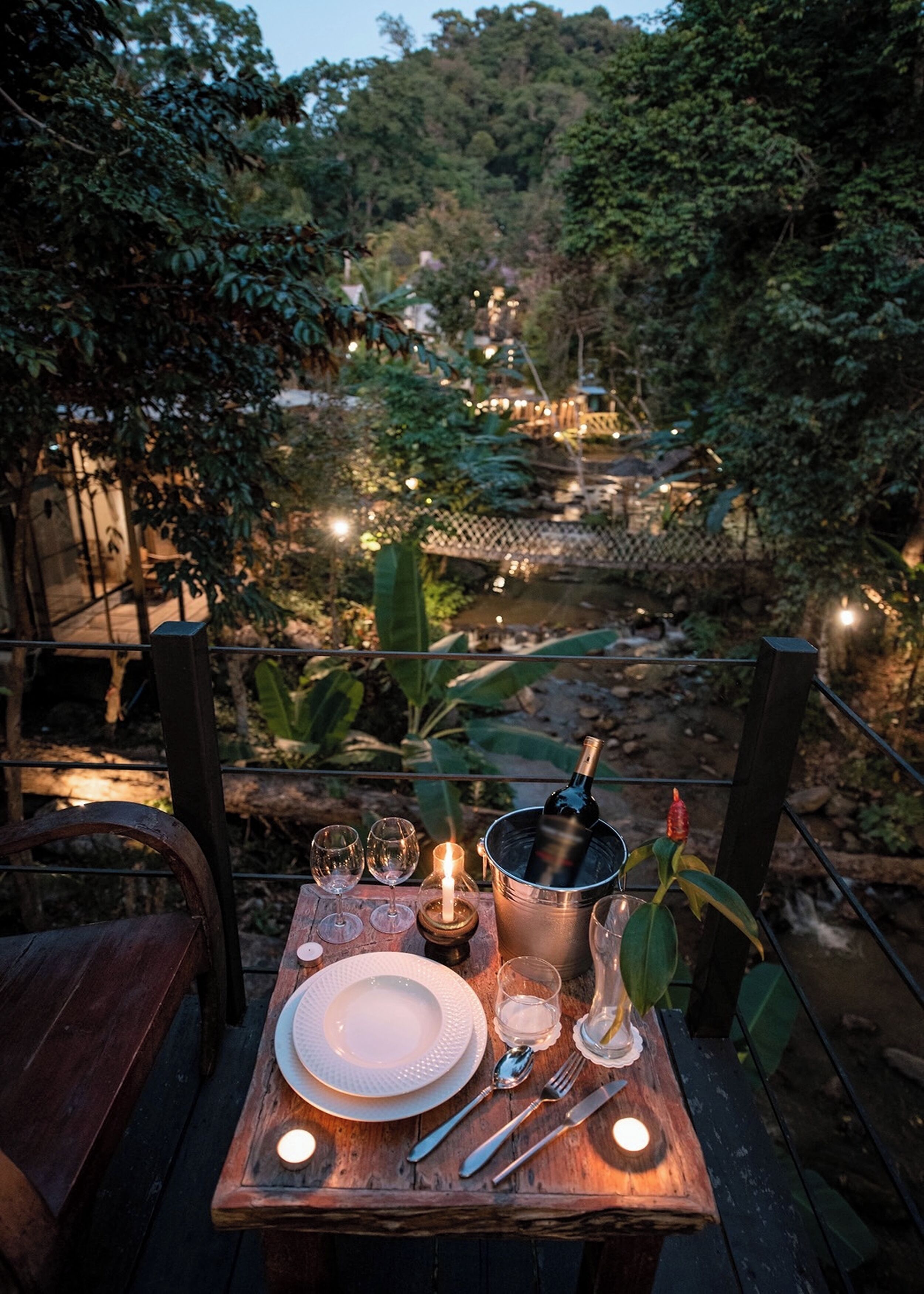 An outdoor dining setup with a table set for two, featuring a bottle of wine, glasses, and a candle.