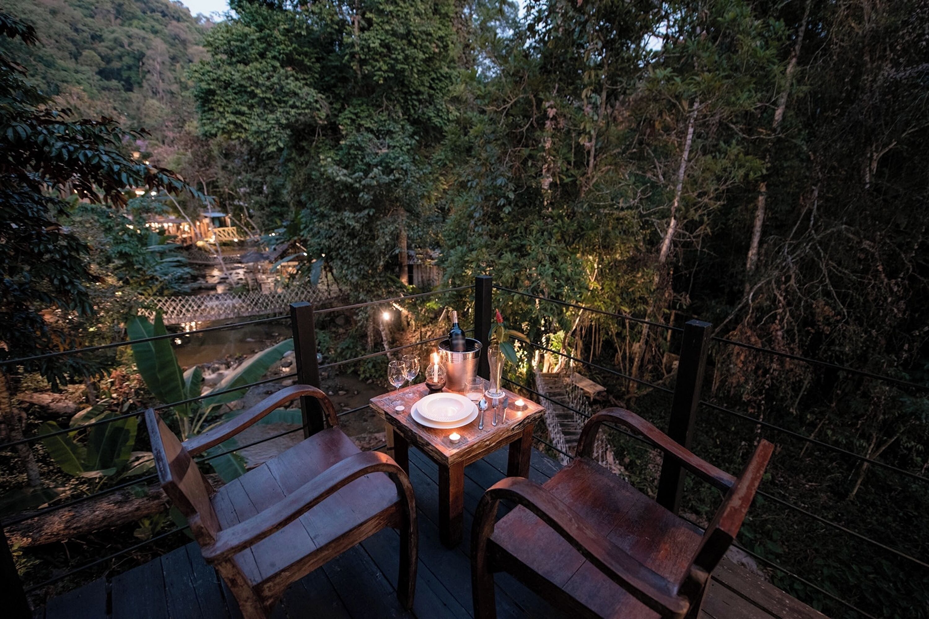 A wooden deck with a table set for two, surrounded by lush greenery and a view of a river.