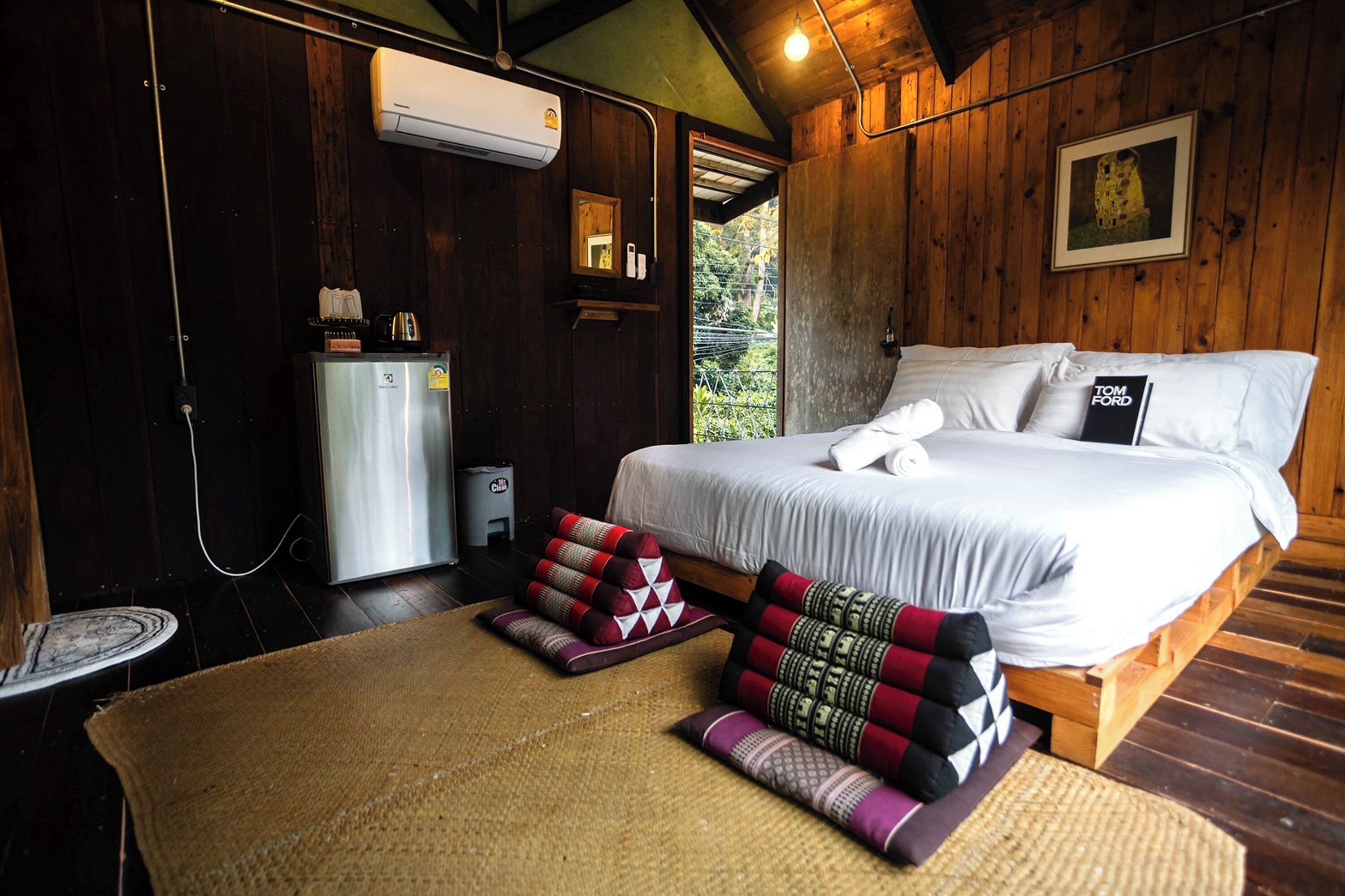 A wooden room with a bed, a small refrigerator, and a window with a view of greenery.