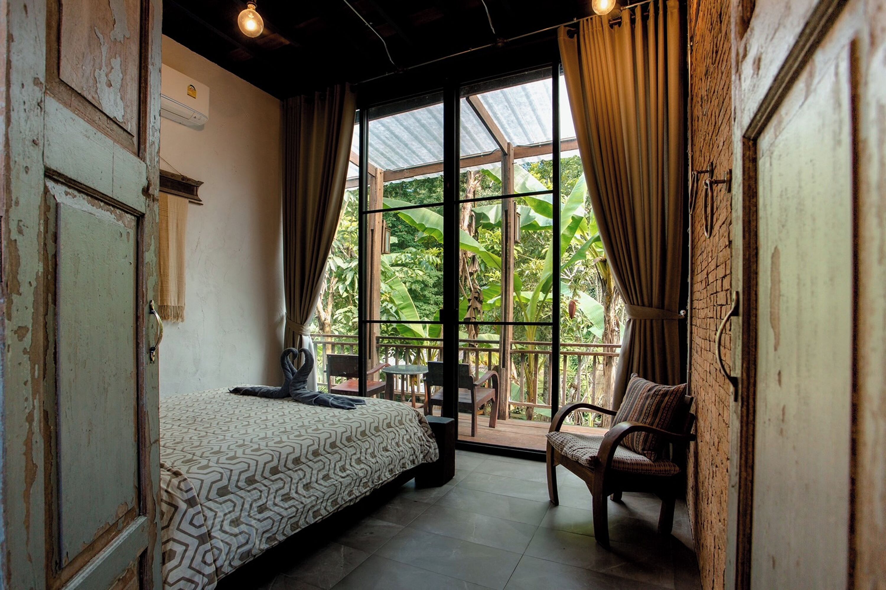 A room with a bed, a chair, and a view of a lush garden through large windows.