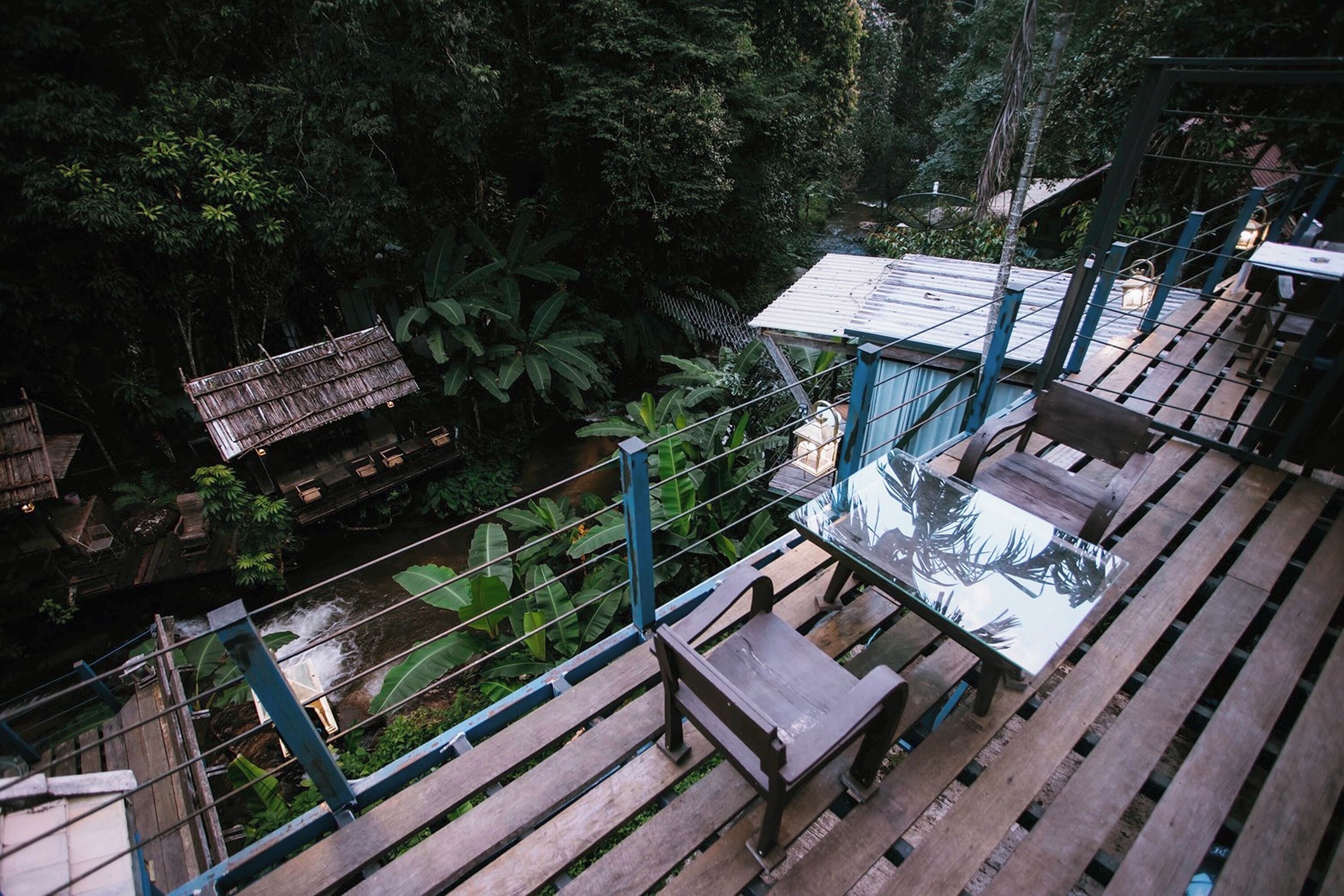 A wooden deck with a glass table and bench, surrounded by lush greenery and a small stream.