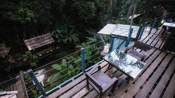 A wooden deck with a glass table and bench, surrounded by lush greenery and a small stream.