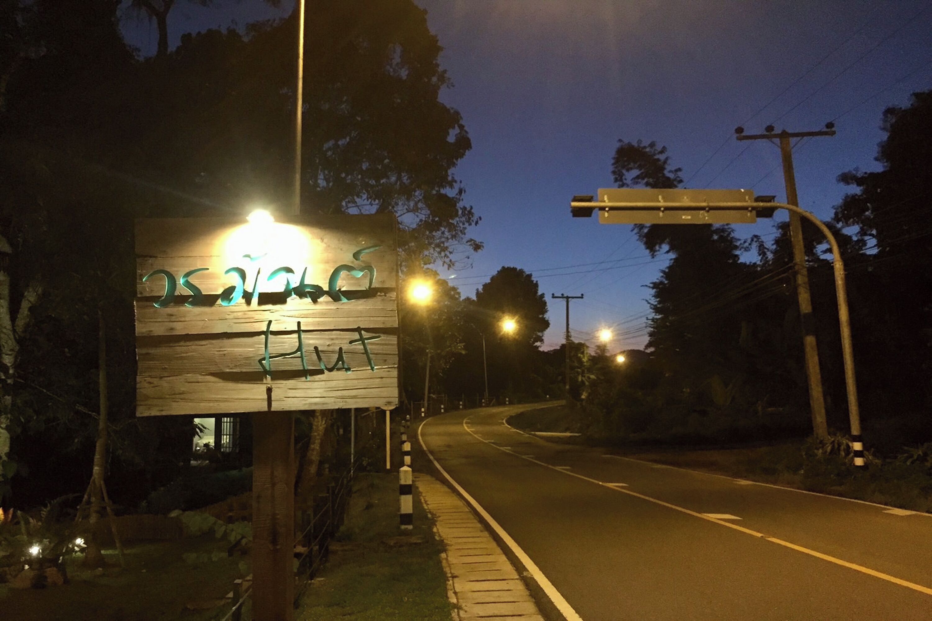 A nighttime street scene with a wooden signboard, streetlights, and a curved road.