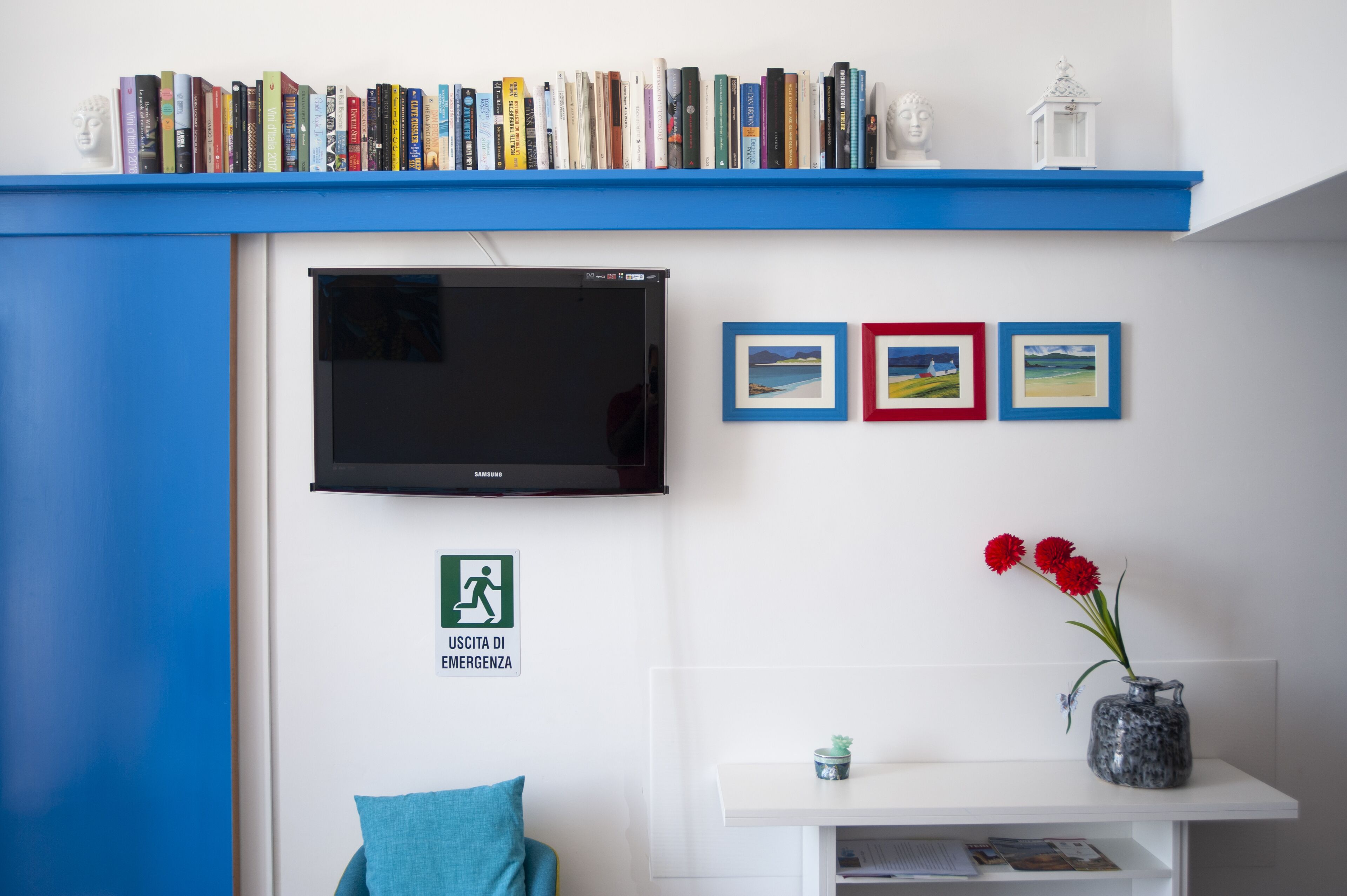 A room with a bookshelf, a flat-screen TV, framed pictures, a vase with flowers, and an emergency exit sign.