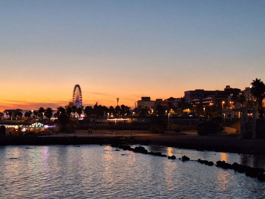 Une vue sur la ville au bord de l’eau, au crépuscule, avec une grande roue bien visible, des bâtiments et une rivière paisible.
