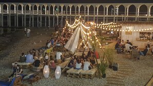 An outdoor gathering with people seated on benches and chairs, a tent with string lights, and a building with arches in the background. - Mowita seaside flat with stunning view on the sea (Civitavecchia)