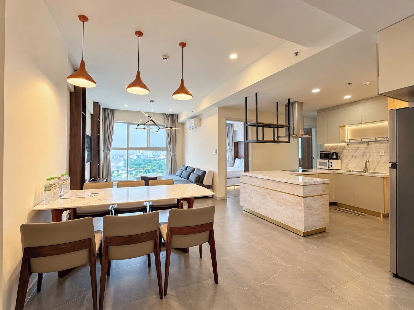 A modern hotel room with a dining area, kitchen, and a large window offering a city view.