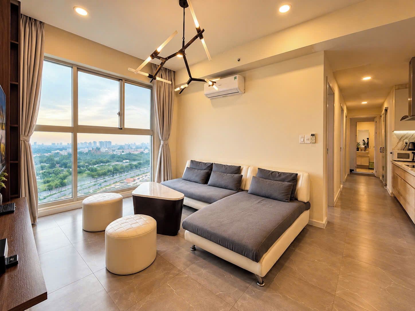 A modern bedroom with a sofa bed, a small round ottoman, a desk with a chair, and a large window offering a city view.