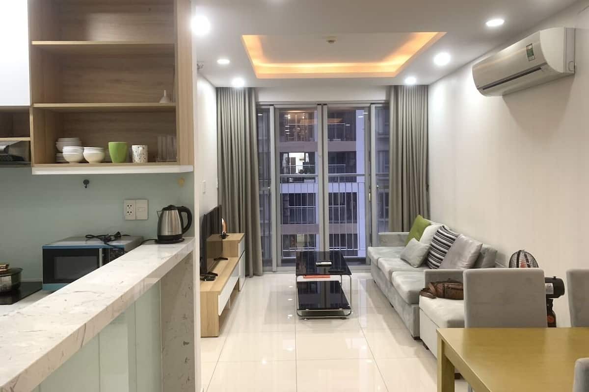 A modern living room with a sofa, a coffee table, and a TV. There is a kitchen area with a microwave and a kettle. The room has a large window with curtains.