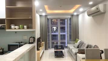 A modern living room with a sofa, a coffee table, and a TV. There is a kitchen area with a microwave and a kettle. The room has a large window with curtains.