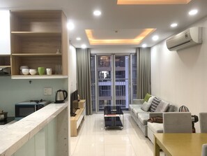 Una sala de estar moderna con un sofá, una mesa de centro y un televisor. Hay una zona de cocina con un microondas y un hervidor. La habitación tiene un ventanal con cortinas.