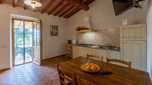 A kitchen with wooden beams, a dining table with chairs, a TV mounted on the wall, and a view through an open door to an outdoor area. - Apartment I Reucci Trilo2 (Pomarance)