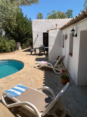Un espace aménagé au bord de la piscine, avec des chaises longues, une table et un petit bâtiment à l’extérieur blanc.