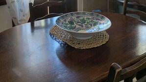 Una mesa de comedor con un cuenco decorativo, sillas de madera, una ventana con contraventanas abiertas y vistas al exterior.