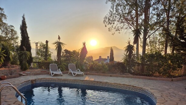 Una piscina con dos sillas reclinables, rodeada de plantas tropicales y vista al atardecer.