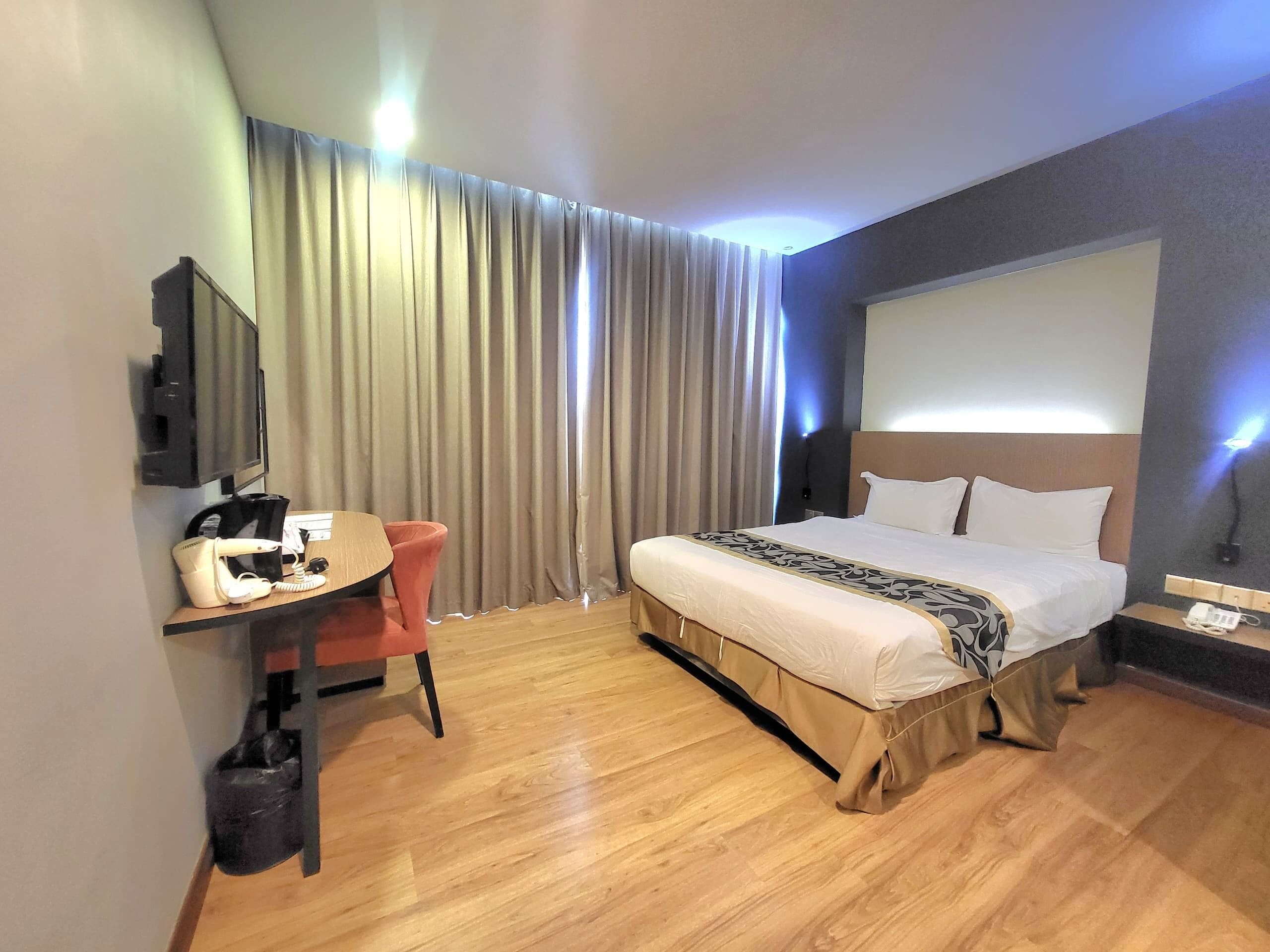 Quarto de hotel com cama, escrivaninha com cadeira, televisão e uma janela grande com cortinas.