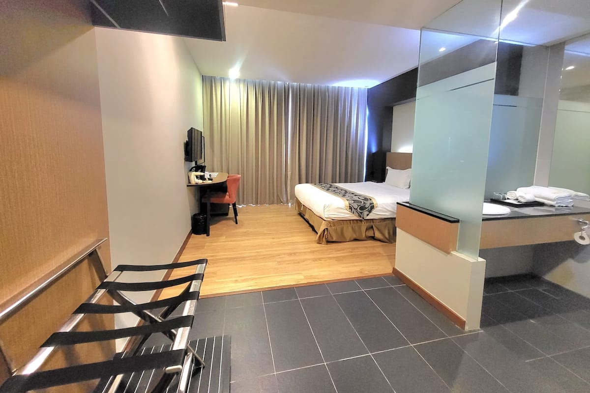 A hotel room with a bed, a desk with a chair, a TV, a mirror, and a bathroom with a towel rack.