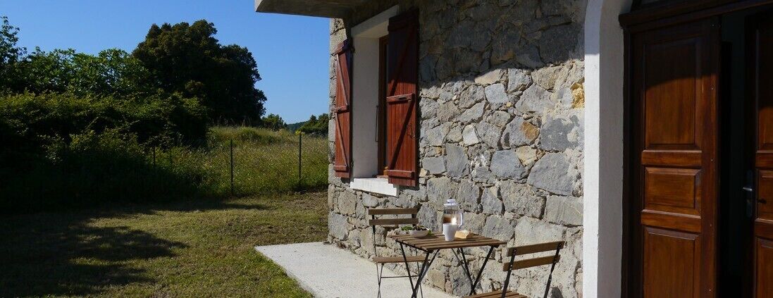 A stone building with a wooden door and window shutters, a small table with chairs, and a lantern on the table.