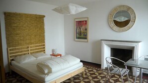 A bedroom with a bed, a painting, a round mirror, a fireplace, and a dining area with chairs.