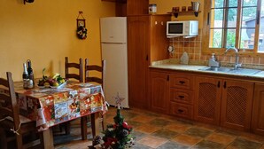 Eine kleine Küche mit Kühlschrank, Mikrowelle und Holzschränken. Im Vordergrund steht ein Esstisch mit Stühlen, und ein kleiner Weihnachtsbaum steht auf einem Couchtisch.