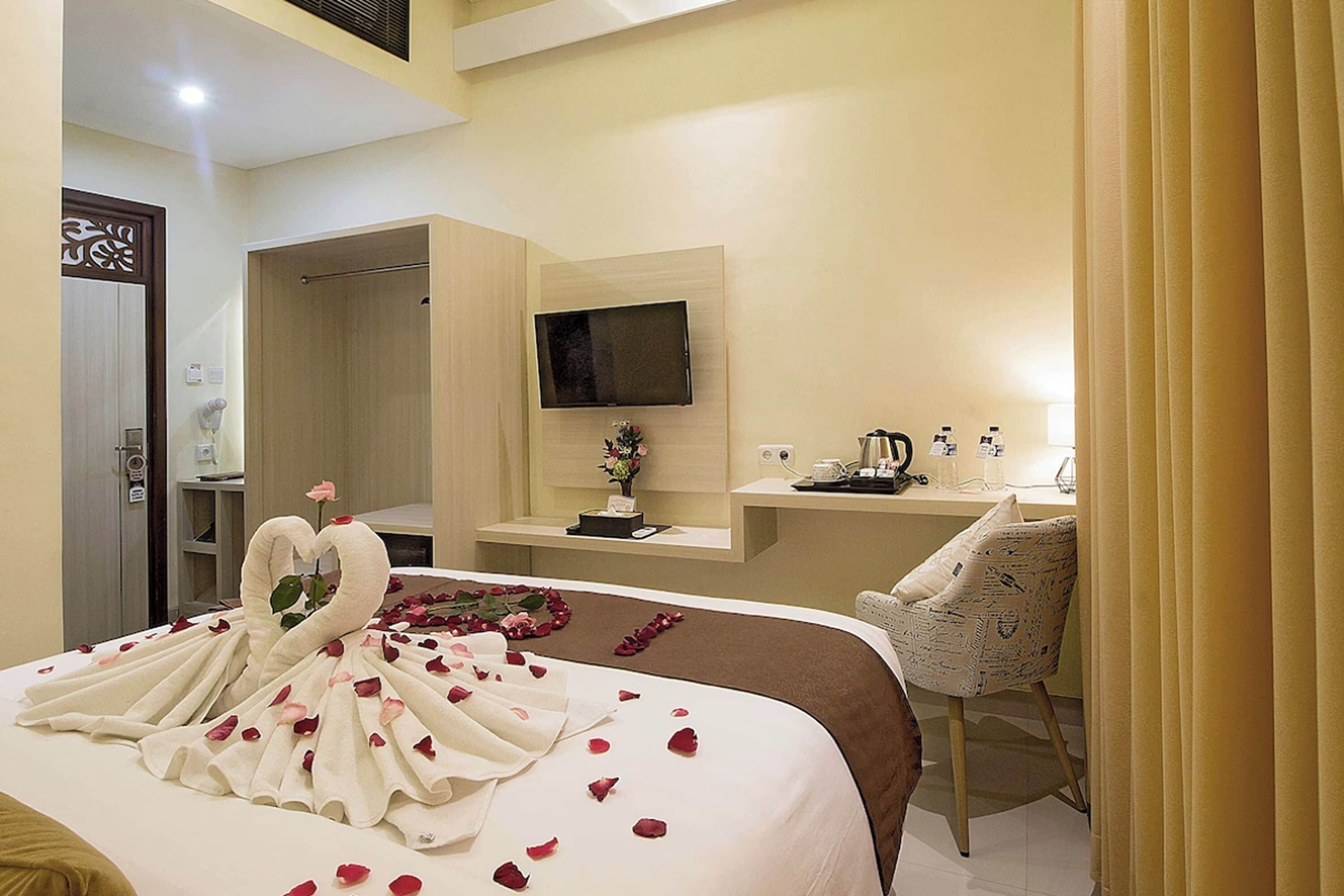 A hotel room with a bed adorned with a swan-shaped towel arrangement, a desk with a computer and a vase of flowers, and a television mounted on the wall.
