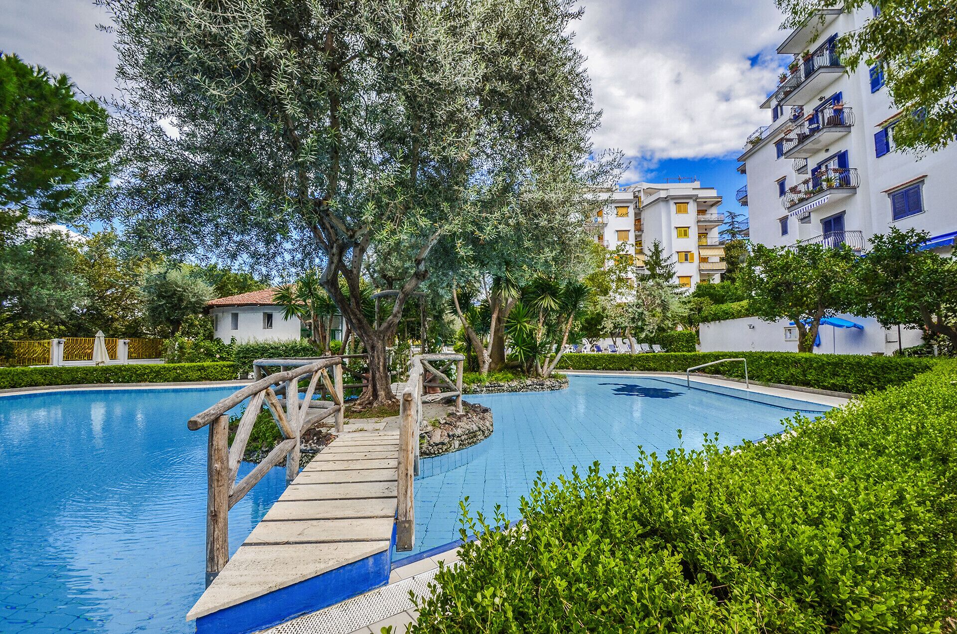 Una piscina con un puente de madera, rodeada de vegetación y edificios residenciales.